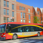 A bright red and yellow DC Circulator bus drives past a tan and orange brick building in Washington, DC