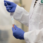 A lab technician wearing a white lab coat with a Puritan logo and blue gloves holds a sterile swab and a vial containing viral transport media in a laboratory setting.