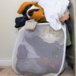 A white mesh laundry basket is leaned up against a dresser. The basket is overflowing with clothes that need to be washed before they’re packed away for a move.
